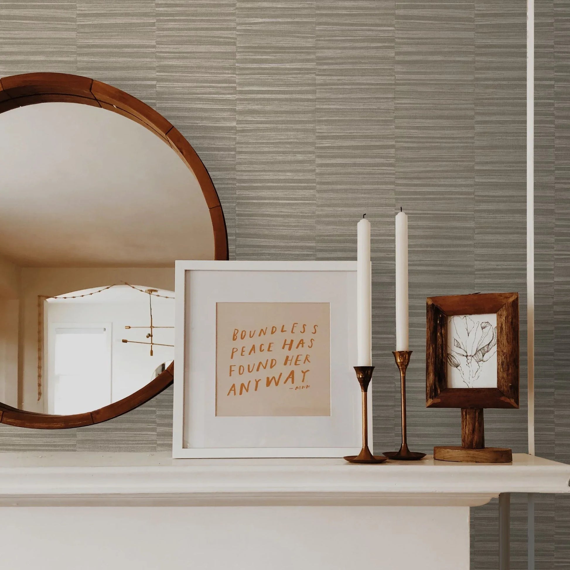 A round mirror and framed art sit on a white mantel against Industrial Grasscloth Stripe vinyl wallpaper.