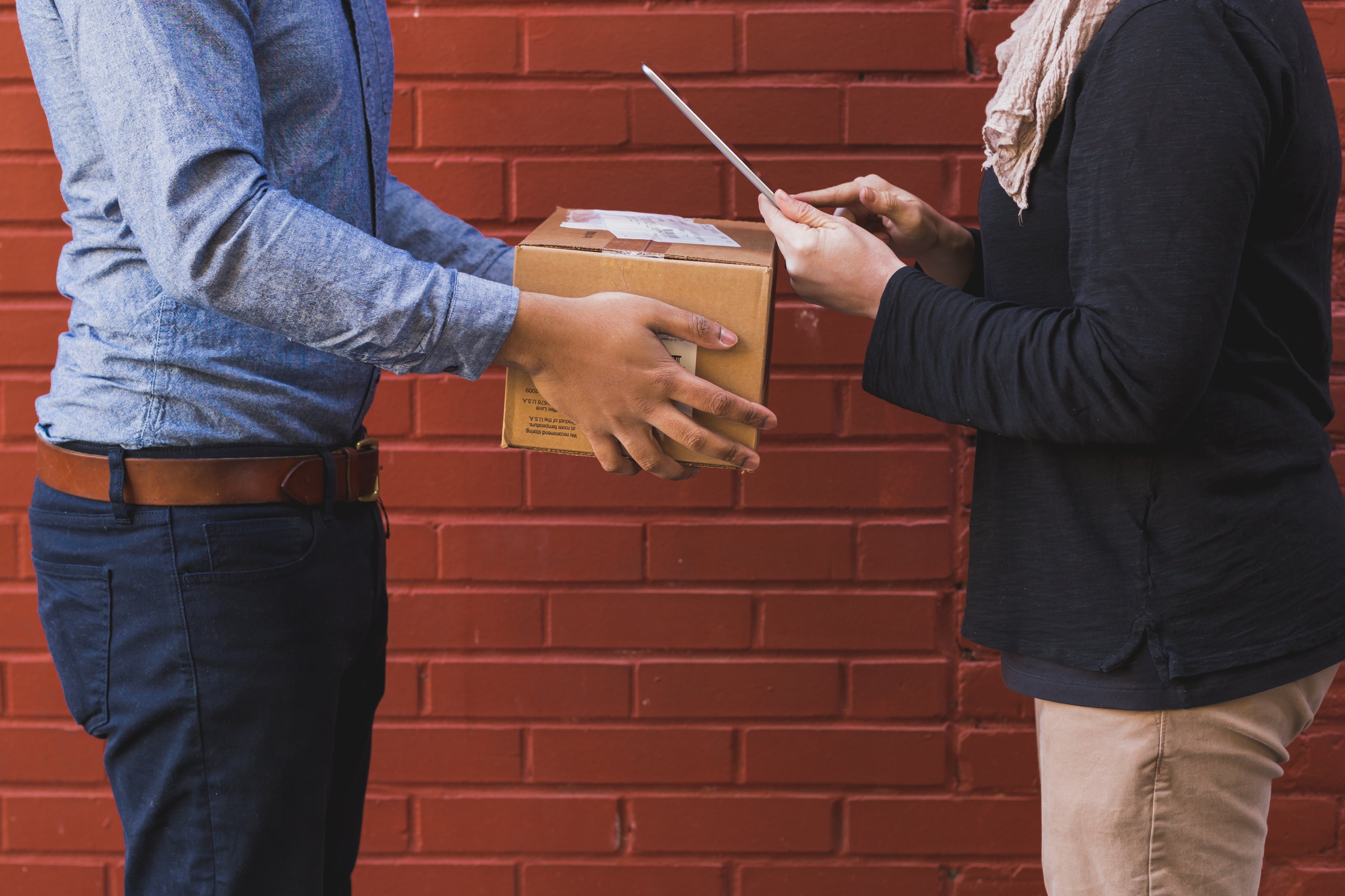 Two people exchange a package and clipboard in front of a red brick wall.
